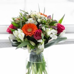 Captivating and expertly composed, this arrangement from a Barnet florist blends radiant colours, sumptuous textures, and local character. Creamy beige and peach gerbera daisies encircle the heart of the bouquet, their gentle petals sculpted in a halo around glowing, orange centers reminiscent of lanterns twinkling in Barnet's evening gardens. Adding a luxurious pop of colour, deep magenta roses spiral upward, each bloom exuding timeless elegance. Between these focal flowers nestle pristine white alstroemeria and sprigs of petite, pale pink filler blooms, creating layers of garden-inspired grace. Broad, paddle-shaped aspidistra leaves are wrapped and curved at the base, acting as a verdant shield that grounds the airy design in lush tranquility, much like the green spaces near Hadley Wood. Smaller pointed foliage weaves through the bouquet, providing subtle volume and a sense of movement. A tall clear glass vase holds the stems, showcasing the freshness and vibrance that's become synonymous with Barnet's local flower deliveries. Soft, ambient light evokes the gentle glow of a north London morning, bathing each petal in natural warmth. This bouquet feels at once celebratory and restorative-suited for gatherings with friends, milestone events, or as a thoughtful gesture to brighten a home near New Barnet Station. Every detail reflects local expertise, care, and a spirit of optimism.